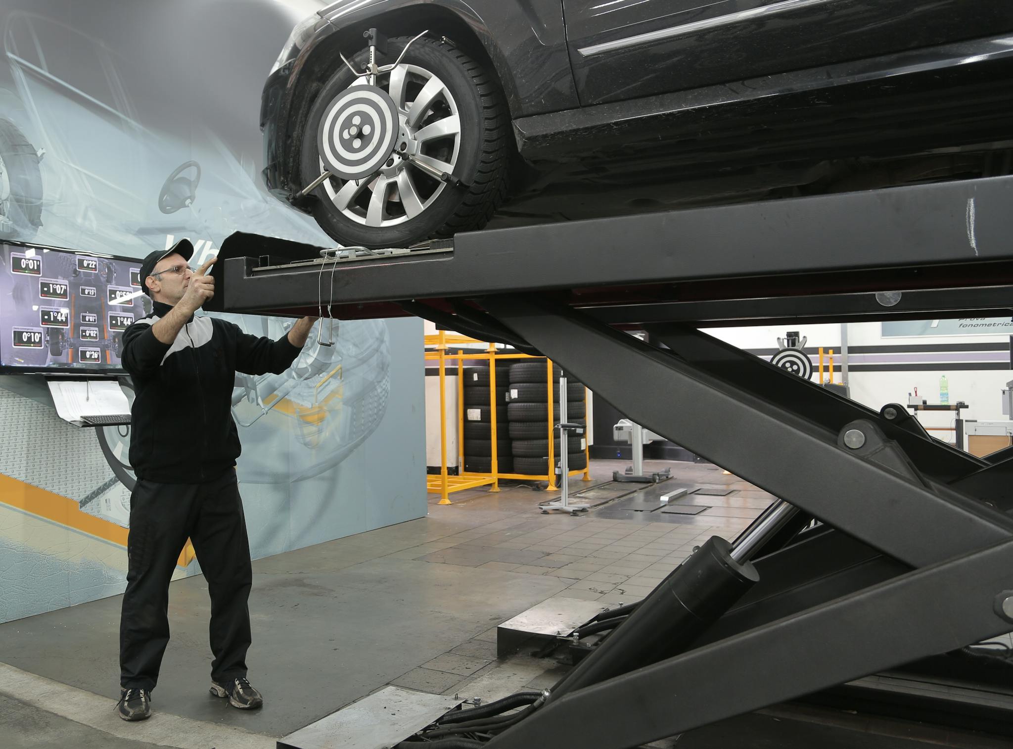 Serious professional adult mechanic in casual uniform examining car wheels while standing near lift in modern car service garage